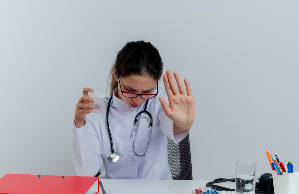 joven-doctora-disgustada-bata-medica-estetoscopio-gafas-sentado-escritorio-herramientas-medicas-sosteniendo-vaso-medico-mirando-abajo-haciendo-gesto-parada-aislado